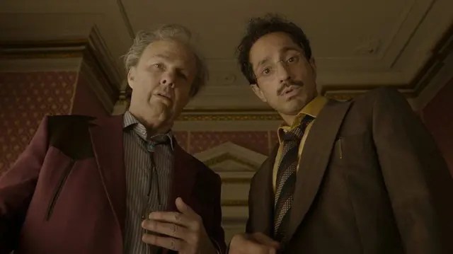 Dos hombres miran hacia abajo con expresiones sorprendidas en un ambiente elegante.