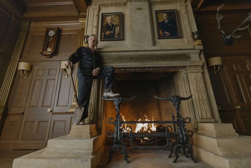 Hombre de pie en una escalera junto a una chimenea grande en una habitación con decoración clásica, incluyendo retratos en la pared.